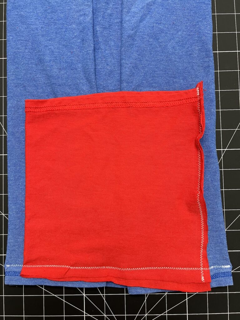 A red knit fabric rectangle with zigzag stitching is sewn onto a blue knit fabric piece, resembling part of a t-shirt bag, both placed on a black grid cutting mat.