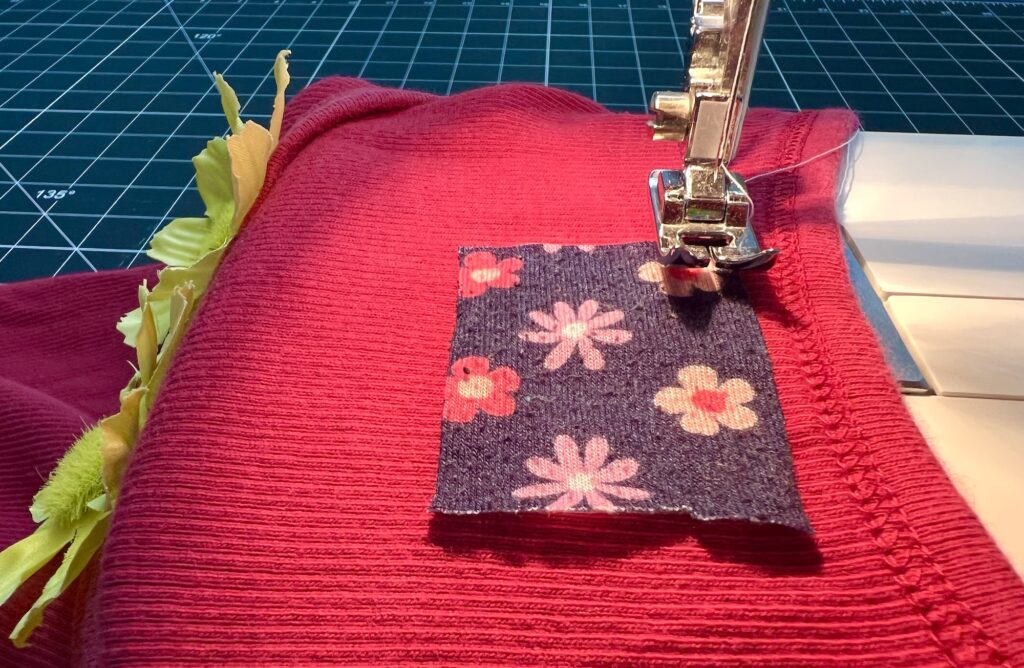 A sewing machine stitches a floral patch onto red fabric, possibly for a t-shirt bag, with a cutting mat visible in the background.