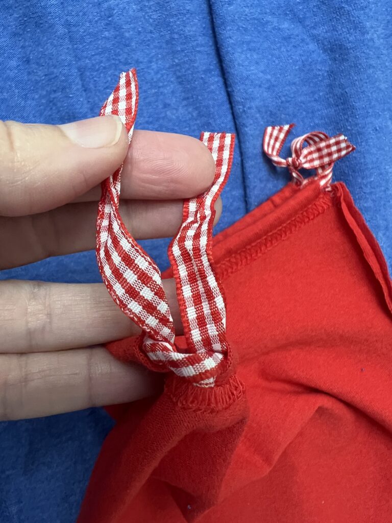 A hand holds two red-and-white checkered ribbons attached to a piece of red t-shirt bag fabric, set against a blue fabric background.