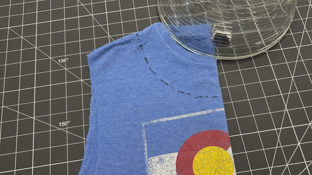 A blue T-shirt is placed on a black grid cutting mat. A glass bowl outlines part of the shirt’s neckline, marked with dashed lines for cutting, as the first step in creating a DIY t-shirt bag.