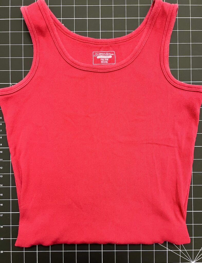 A red ribbed tank top, perfect for upcycling into a t-shirt bag, is laid flat on a black grid-patterned cutting mat.