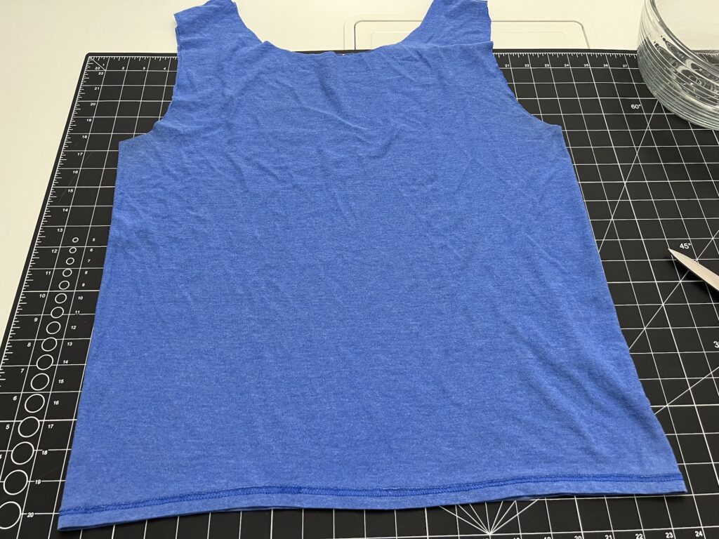A sleeveless blue shirt, ideal for making a t-shirt bag, is made from T-shirt material and laid flat on a black cutting mat with measurement markings.