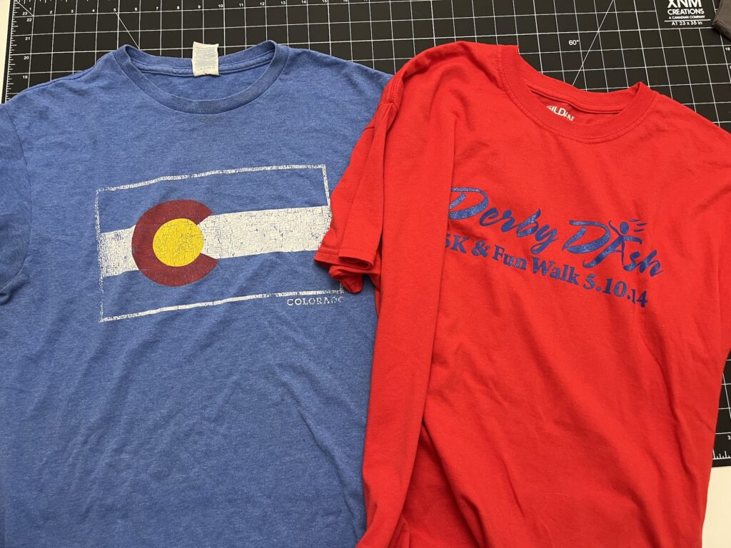A blue Colorado flag T-shirt and a red “Derby Dish 5K & Fun Walk 5.10.14” T-shirt, perfect for making a t-shirt bag, are laid out on a black cutting mat.