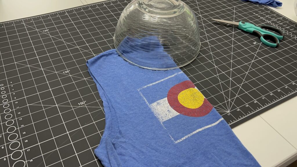 A blue T-shirt with a Colorado flag design is laid on a cutting mat, partially covered by an upside-down glass bowl; scissors are nearby, ready for crafting it into a t-shirt bag.