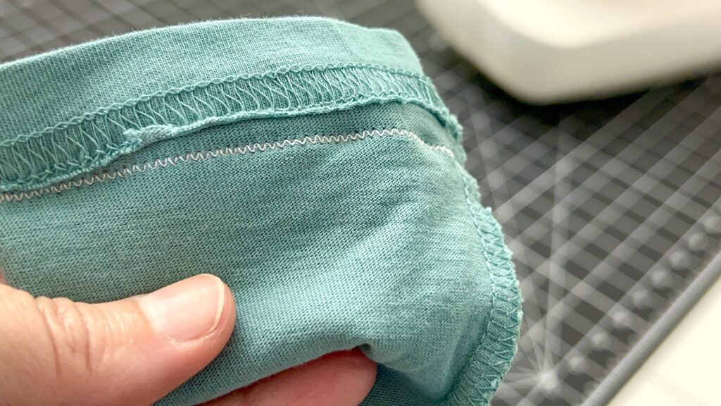 A hand holds a piece of teal knit fabric—soon to become a t-shirt bag—with visible serged seams and zigzag stitching, set on a cutting mat near a sewing machine.