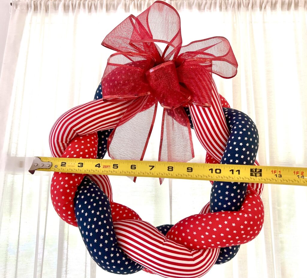A red, white, and blue braided wreath with stars and stripes and a red bow is being measured with a yellow tape measure; it reads about 18 inches in diameter.