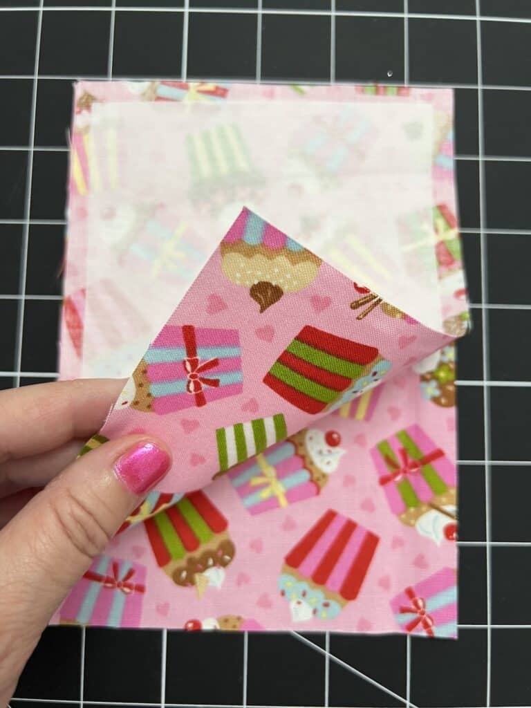A hand with pink nail polish lifts a corner of pink fabric with cupcake prints, revealing interfacing on the back, on a black gridded surface.