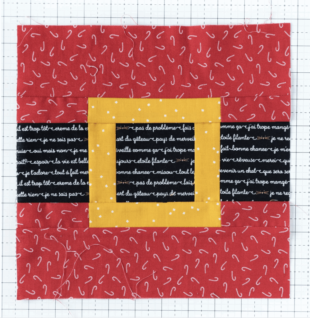 A quilt block with a red background featuring candy cane patterns, a yellow frame, and black strips with white cursive text, laid on grid paper.