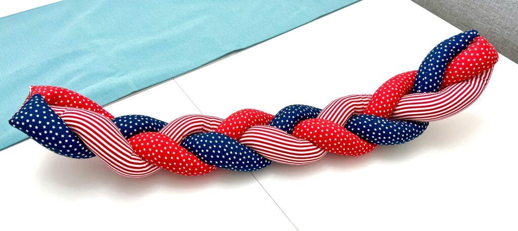 A braided cushion made of red, white, and blue fabric with stripes and stars, placed on a white table near a light blue cloth.