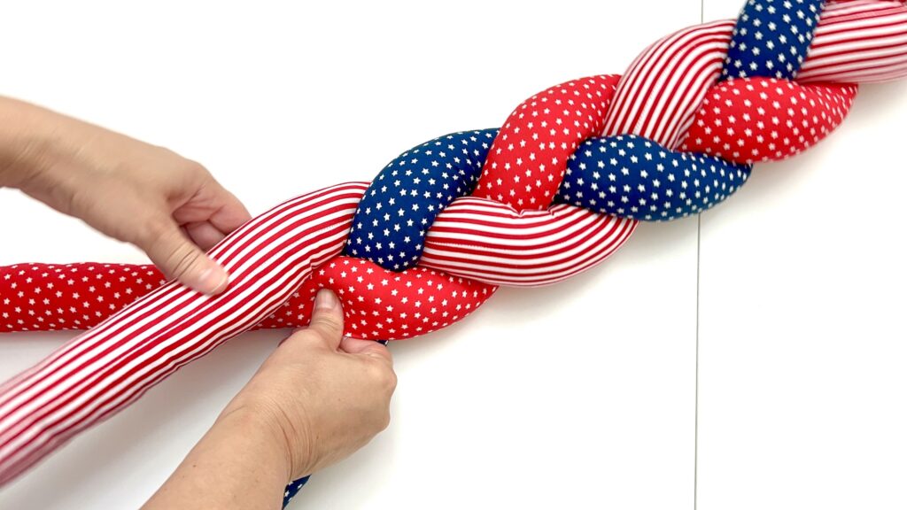 Two hands are braiding a soft, padded fabric in red, white, and blue with star and stripe patterns on a white surface.