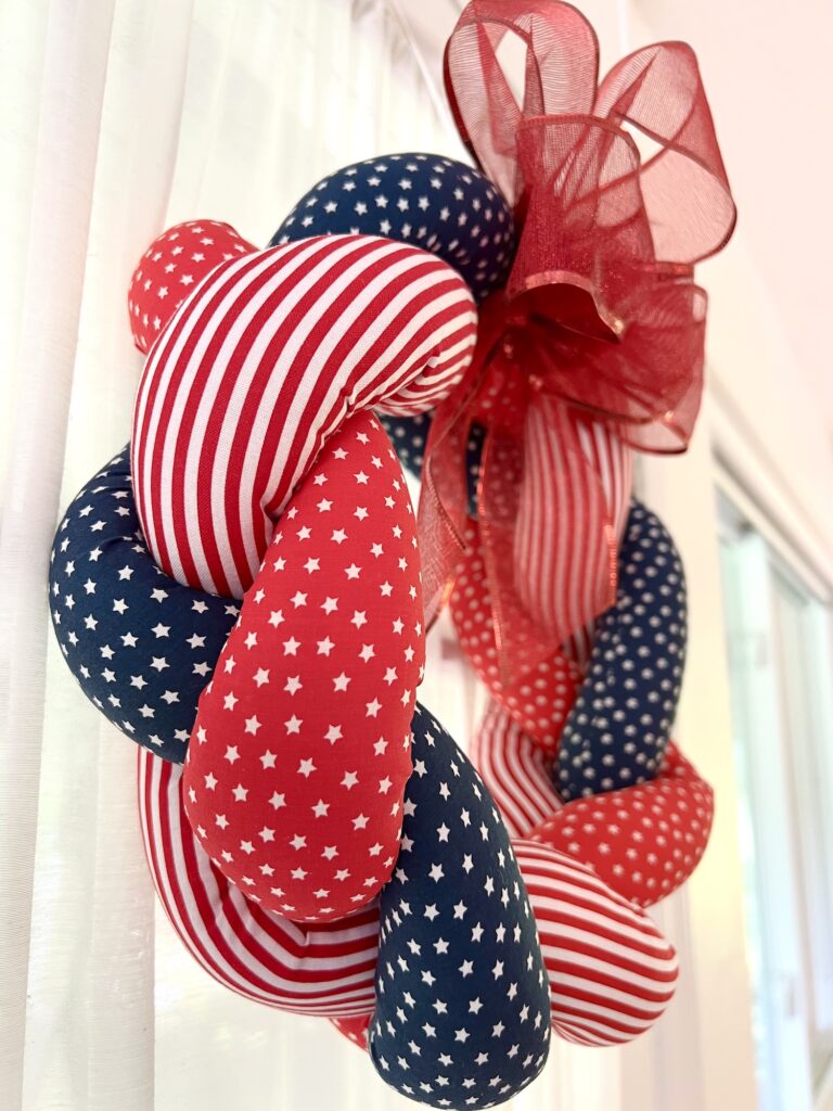 A braided wreath made of red, white, and blue fabric with stars and stripes patterns, adorned with a large red mesh bow, hangs indoors near a window.