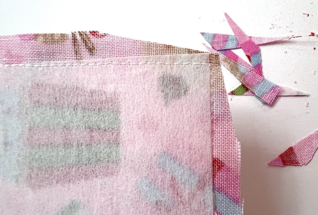 Close-up of a piece of pink patterned fabric with stitched edges, trimmed corners, and fabric scraps cut off lying nearby on a white surface.