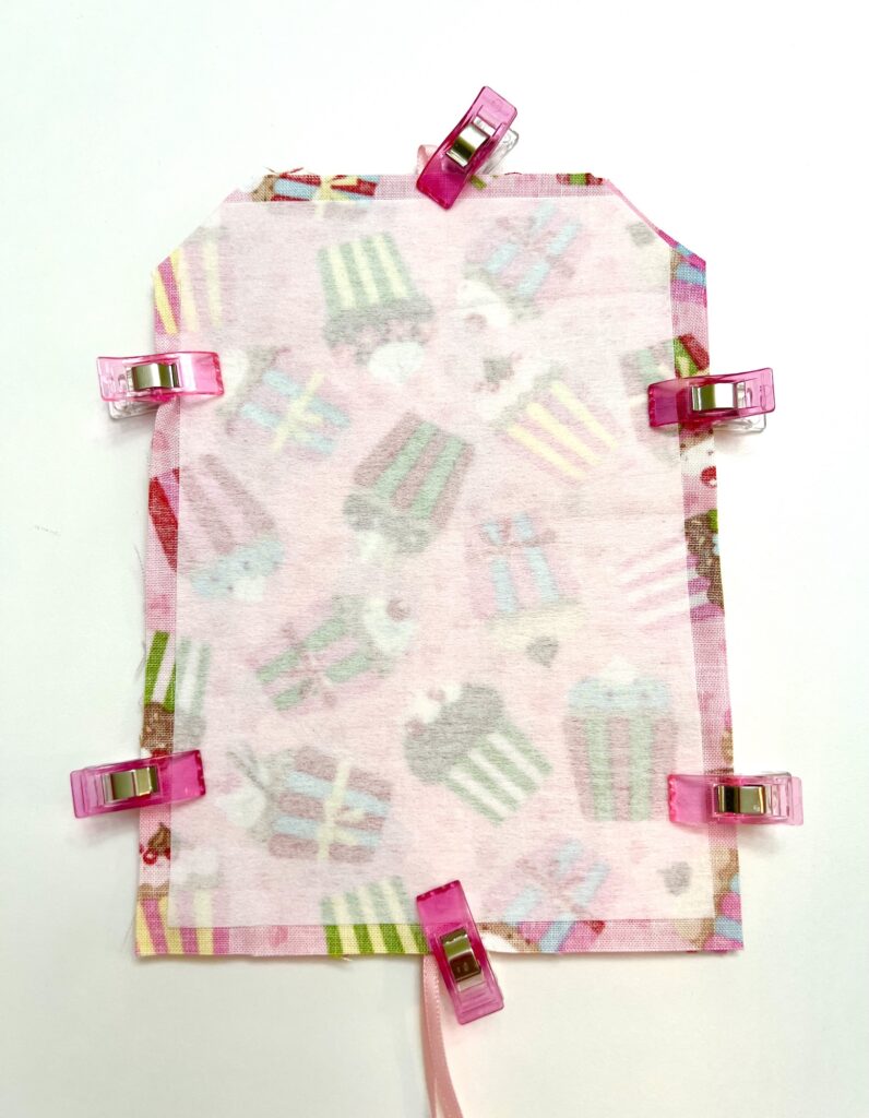Two pieces of patterned fabric are clipped together with six pink sewing clips, ready to be sewn. The fabric features images of cupcakes and gifts.