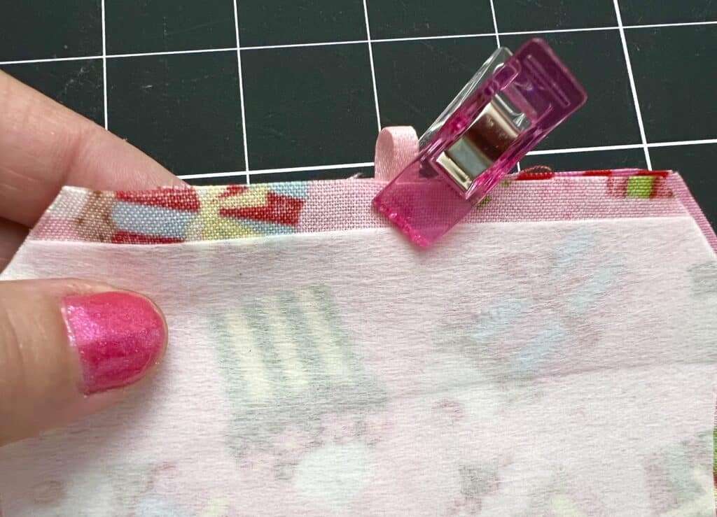 A hand with pink nail polish holds layered pink fabric pieces secured with a pink clip, against a black grid-patterned surface.