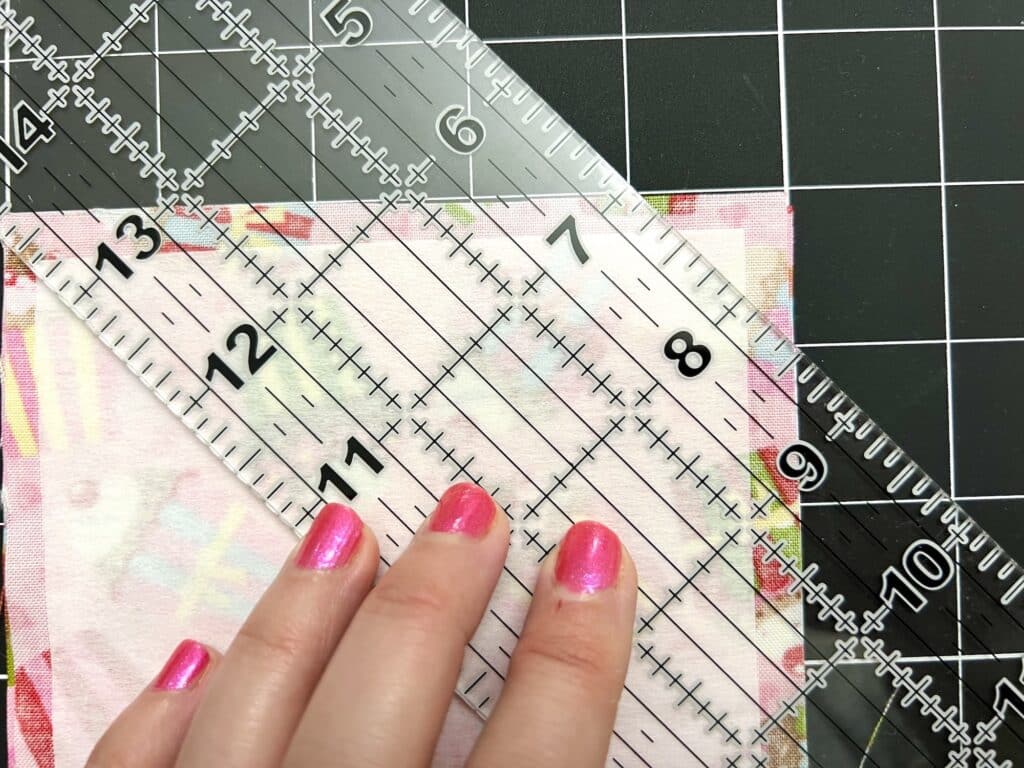 A hand with pink nail polish holds a clear quilting ruler over a piece of fabric on a black cutting mat with white gridlines.