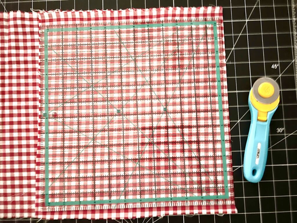 A rotary cutter and a clear ruler are placed on top of a red and white checkered fabric, all set on a black cutting mat with grid lines.