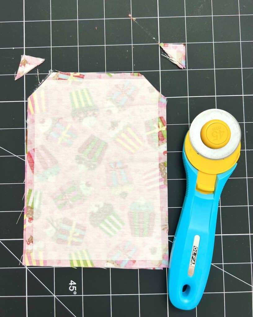 A piece of fabric with trimmed corners lies on a cutting mat next to a blue and yellow rotary cutter.