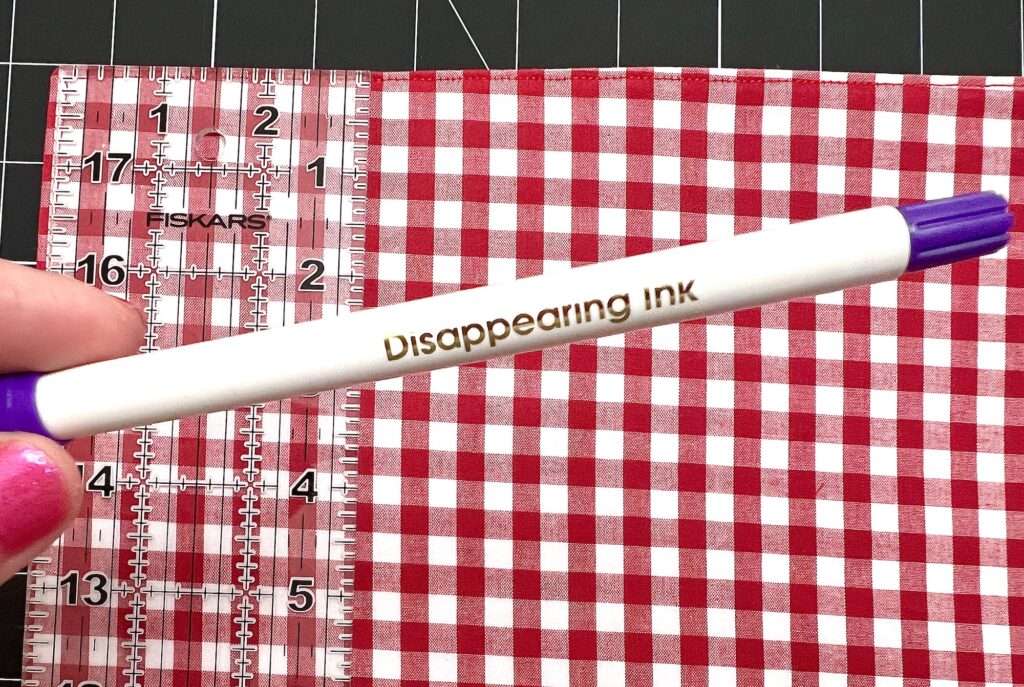 A hand holds a white pen labeled "Disappearing Ink" over a red and white checkered fabric and a clear ruler.