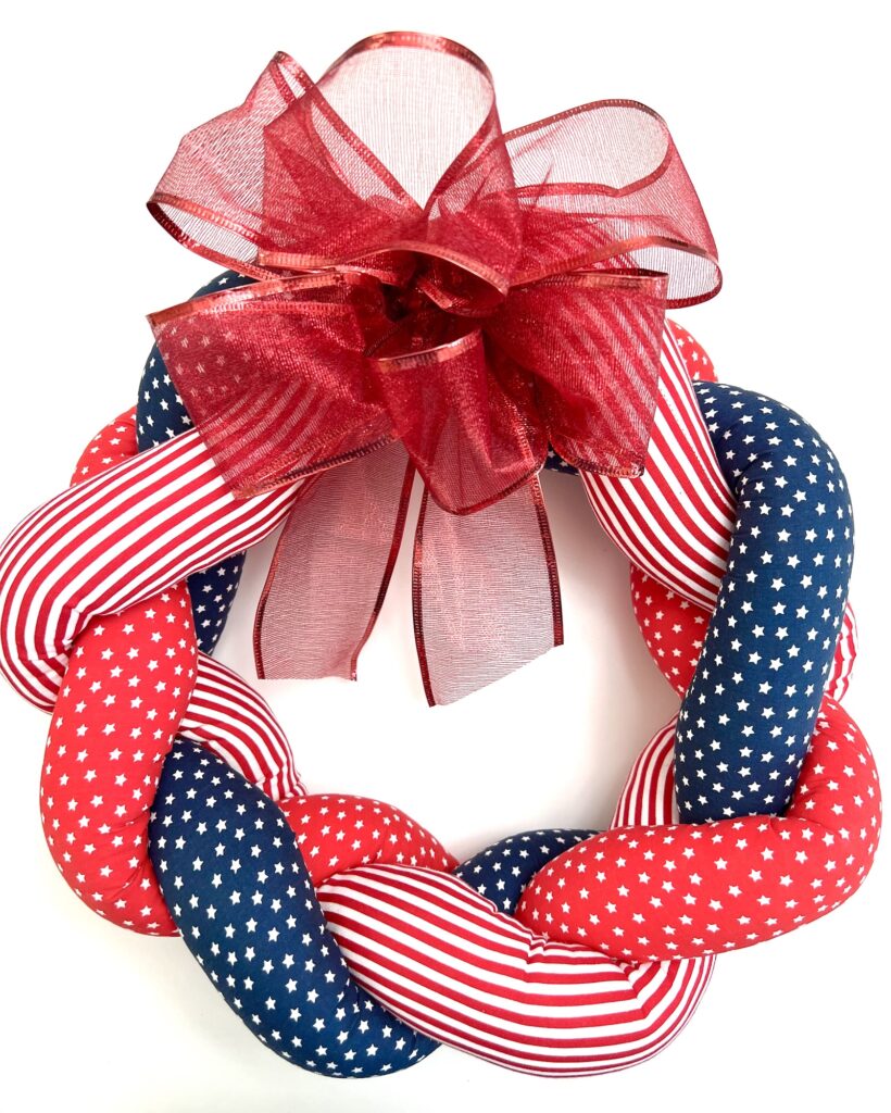 A braided wreath with red, white, and blue fabric featuring stars and stripes, topped with a large red sheer ribbon bow.