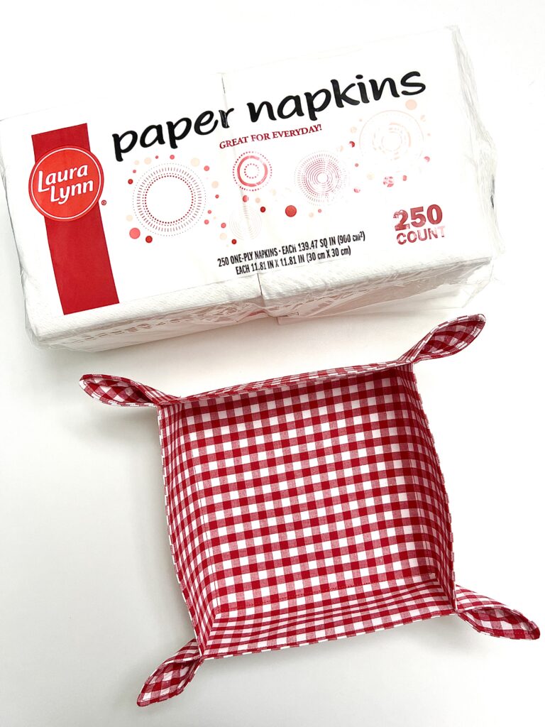 A pack of Laura Lynn paper napkins and a red and white checkered fabric basket are displayed on a white background.