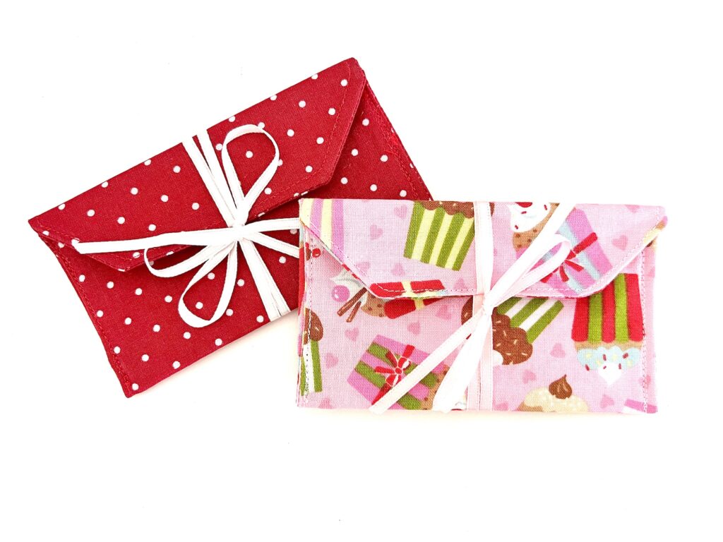 Two fabric envelope pouches, one red with white polka dots and one pink with a dessert pattern, both tied with white ribbon bows.