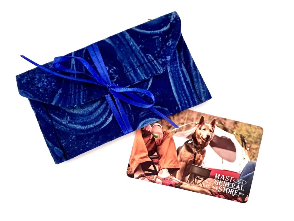 A blue fabric envelope with a ribbon next to a Mast General Store gift card featuring a camping scene with a German Shepherd dog.
