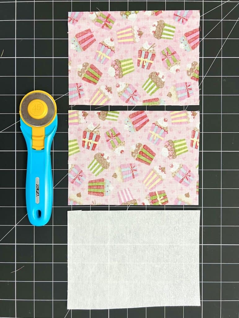Three fabric rectangles with a cupcake pattern, a plain white rectangle, and a blue rotary cutter are arranged on a black cutting mat with a grid.
