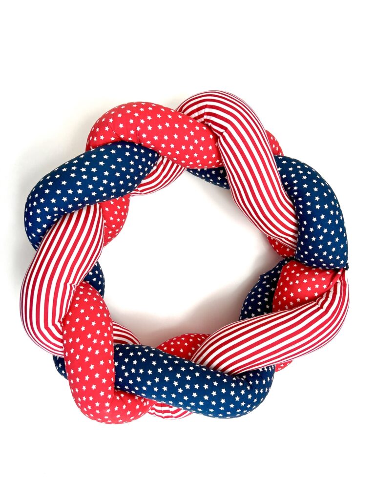 A braided wreath made of red, white, and blue fabric with stars and stripes patterns, arranged in a circular shape on a white background.