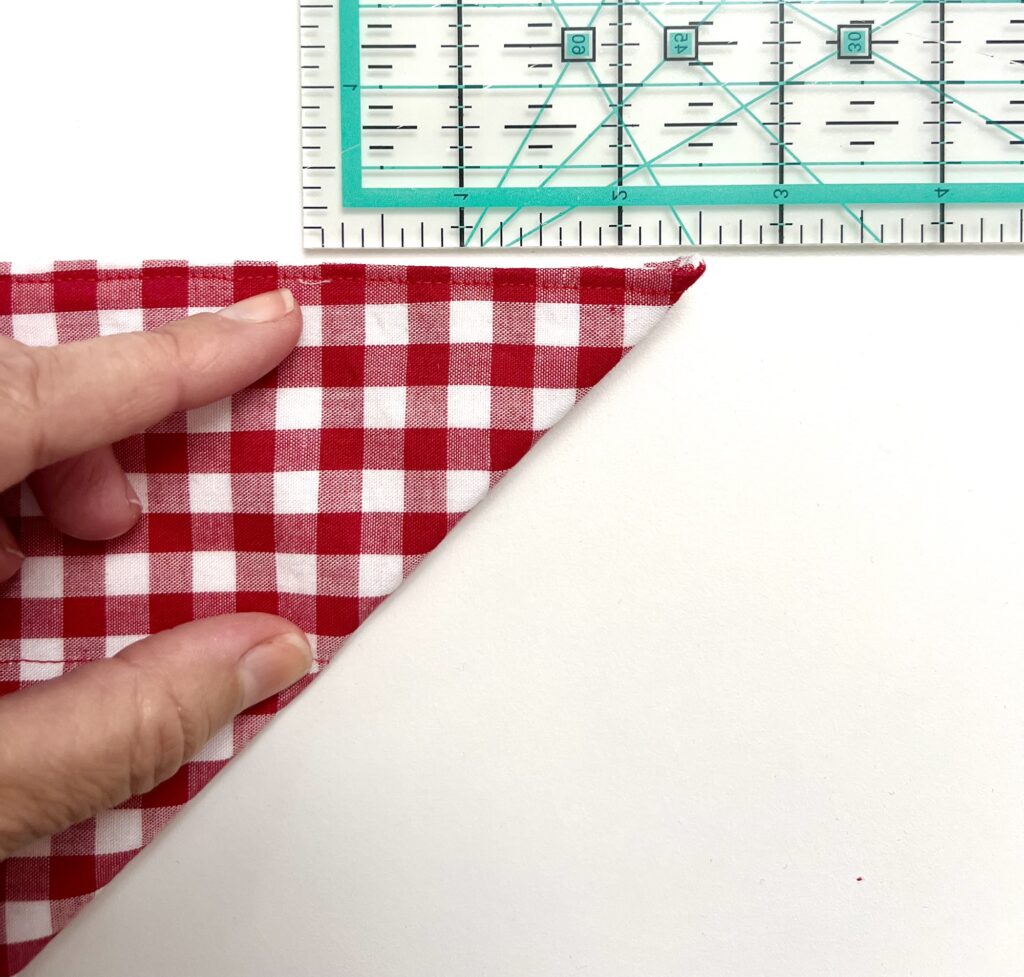 A hand holds the corner of a red and white checkered fabric folded into a triangle next to a clear plastic ruler on a white surface.