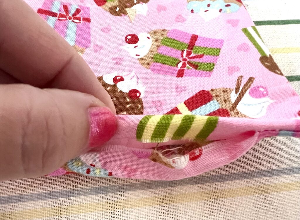 Close-up of a hand with pink nail polish holding the hem of a pink fabric with a cupcake and present pattern, showing loose threads at the seam.