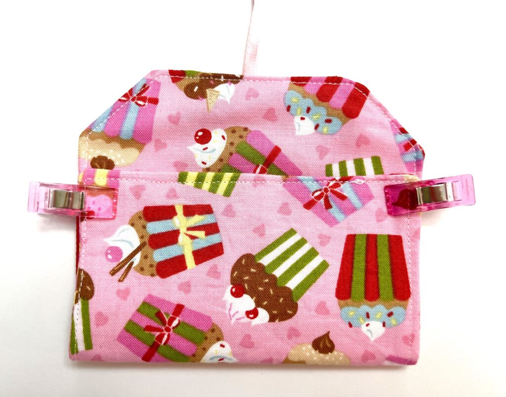 A small pink fabric pouch with a cupcake and gift pattern, clipped at both sides with pink clips, against a white background.