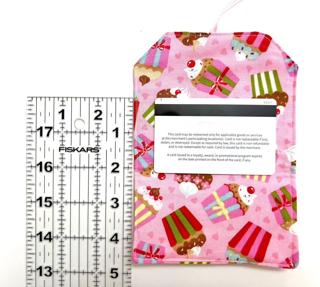 A pink fabric pouch with cupcake and present designs holds a card partially inserted; a ruler beside it shows the pouch is about 6.5 inches long.