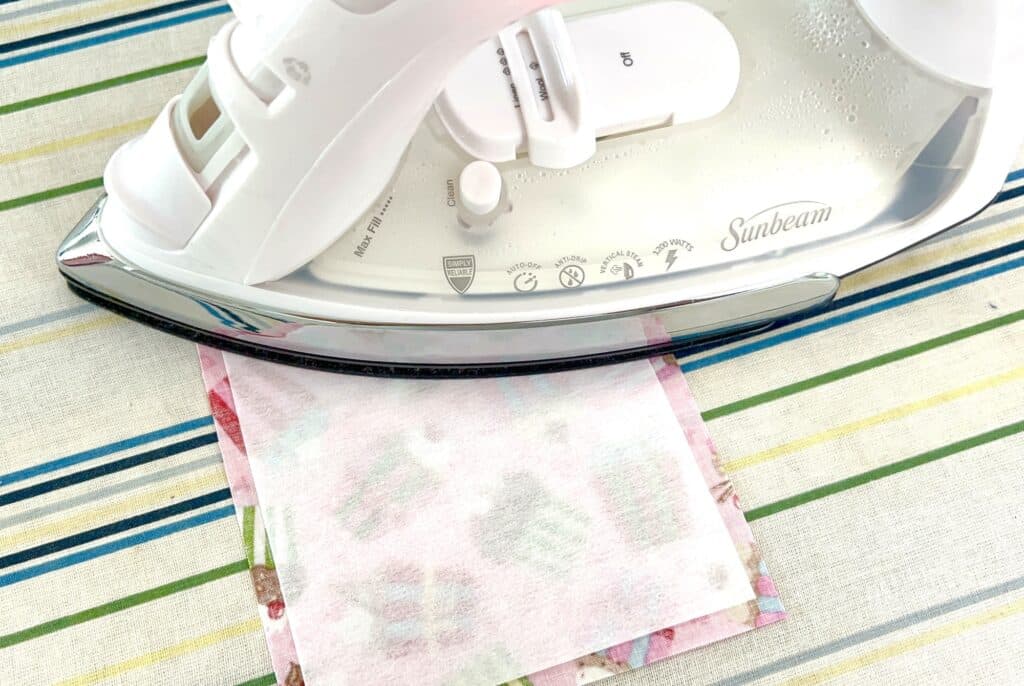 A white iron presses a small square of fabric with a paper backing on a striped ironing board.