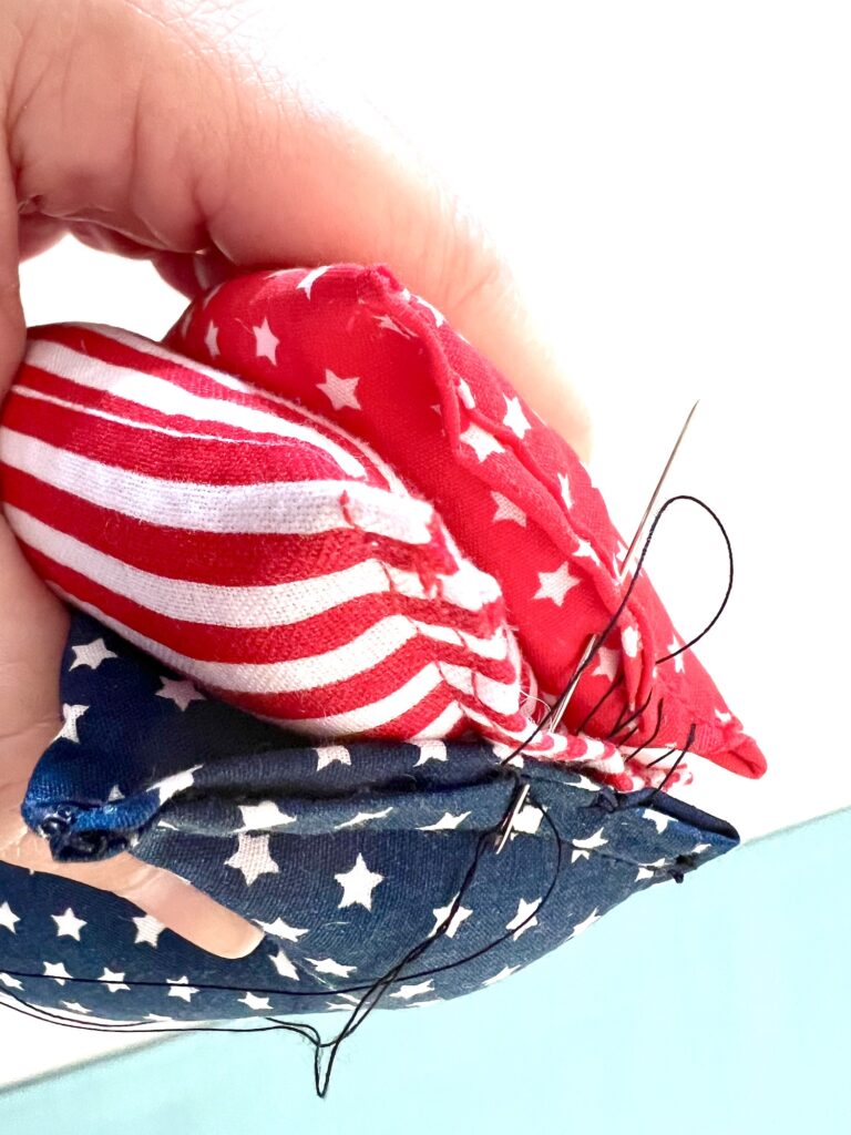 A hand is sewing together pieces of fabric with red stripes and stars, using a needle and black thread. The fabric resembles an American flag pattern.