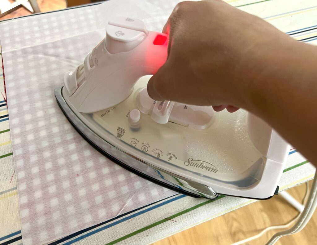 A person ironing a piece of light purple checkered fabric on an ironing board using a white Sunbeam iron.