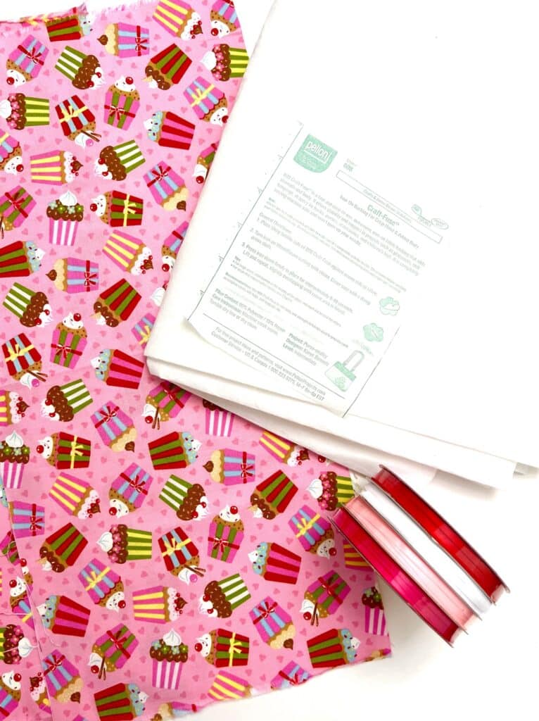 Pink cupcake-patterned fabric, white paper with printed instructions, and three spools of pink and red ribbon are arranged on a white surface.