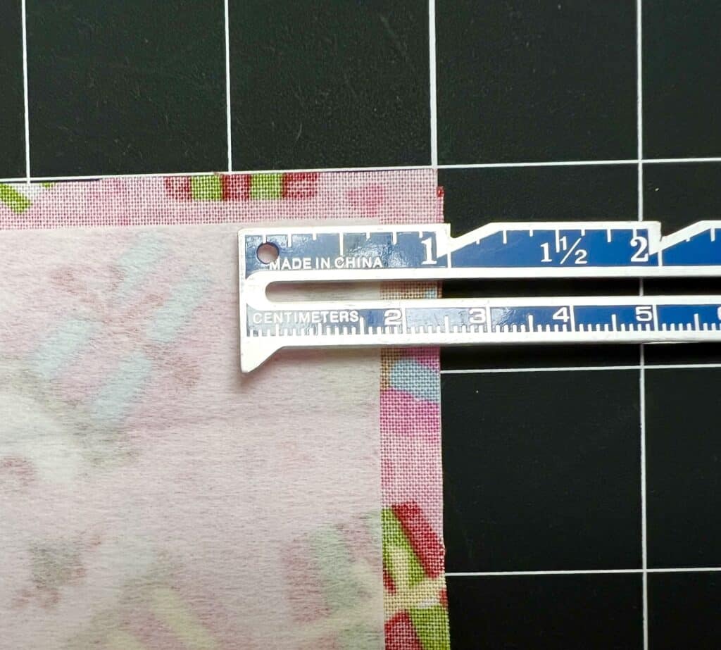 A sewing gauge measuring a 1/2 inch seam allowance on a piece of floral fabric, placed on a black gridded cutting mat.
