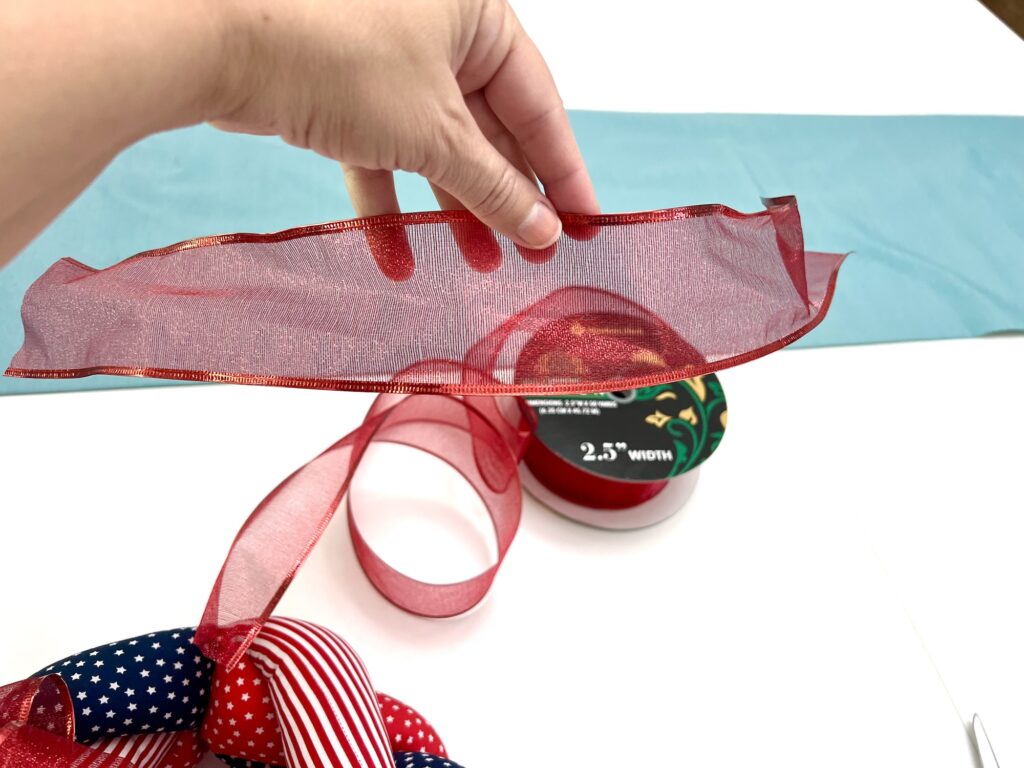 A hand holds a strip of sheer red ribbon above a table with a spool of ribbon, a blue fabric strip, and patriotic-themed ribbon.