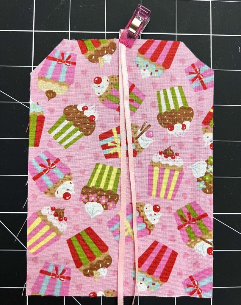 A piece of pink fabric with a cupcake and gift box pattern, clipped at the top with a pink clip, lying on a black grid cutting mat.
