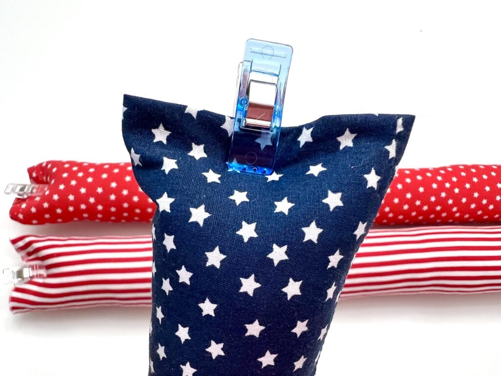 A blue fabric pouch with white stars is clipped shut on top, in front of two red and white patterned fabric rolls on a white background.