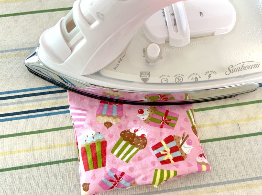 A white steam iron pressing a small pink fabric pouch decorated with cupcakes and gifts on a striped ironing board.