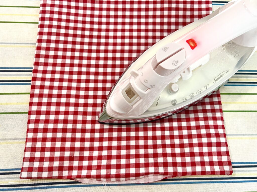 A white iron with a red light is pressing a red and white checkered cloth on a striped ironing board cover.