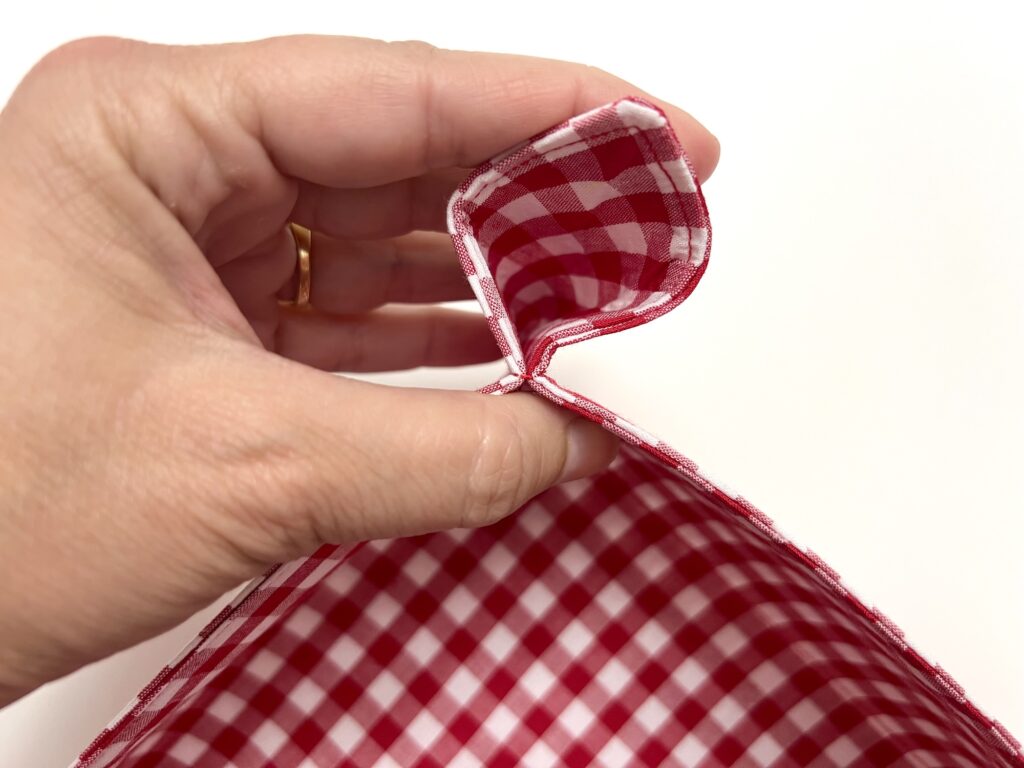 A hand holding the corner of a red and white checkered fabric item, showing its sewn edge and interior pattern against a white background.
