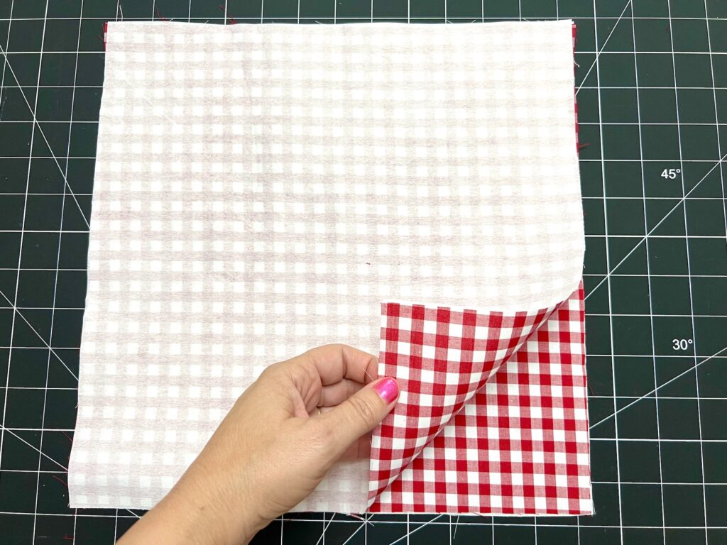 A hand with pink nail polish lifts the corner of a red and white checkered fabric, revealing a lighter checkered fabric beneath, on a cutting mat.