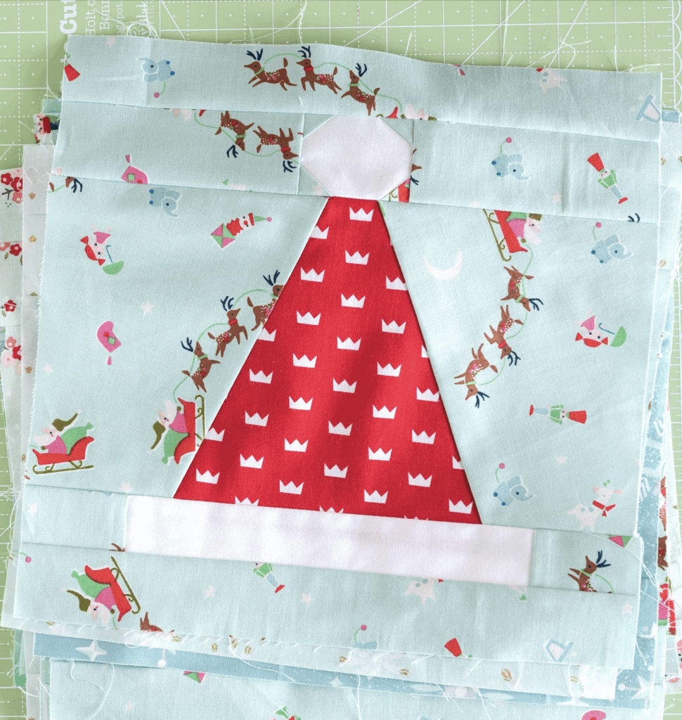 A quilt block featuring a red Santa hat with white trim, surrounded by light blue fabric with festive prints of reindeer, sleighs, and gifts.