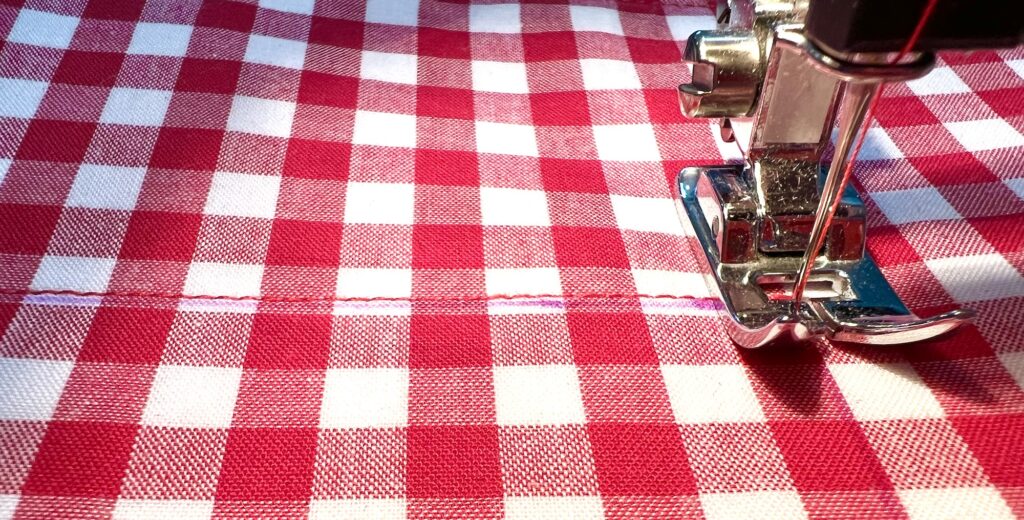 A close-up of a sewing machine stitching a straight line on red and white checkered fabric.