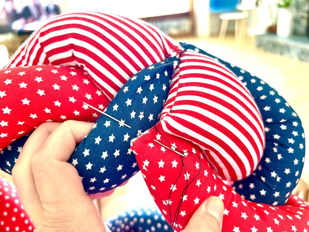 A hand sewing together red and blue fabric pieces with white stars and stripes, using a needle and thread.
