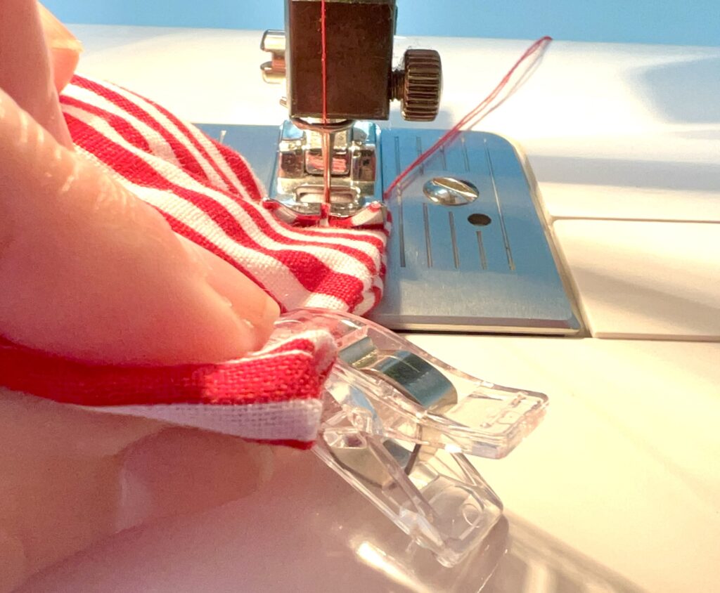 Close-up of a person's hand guiding red and white striped fabric under the needle of a sewing machine, with a clear plastic clip holding the fabric in place.