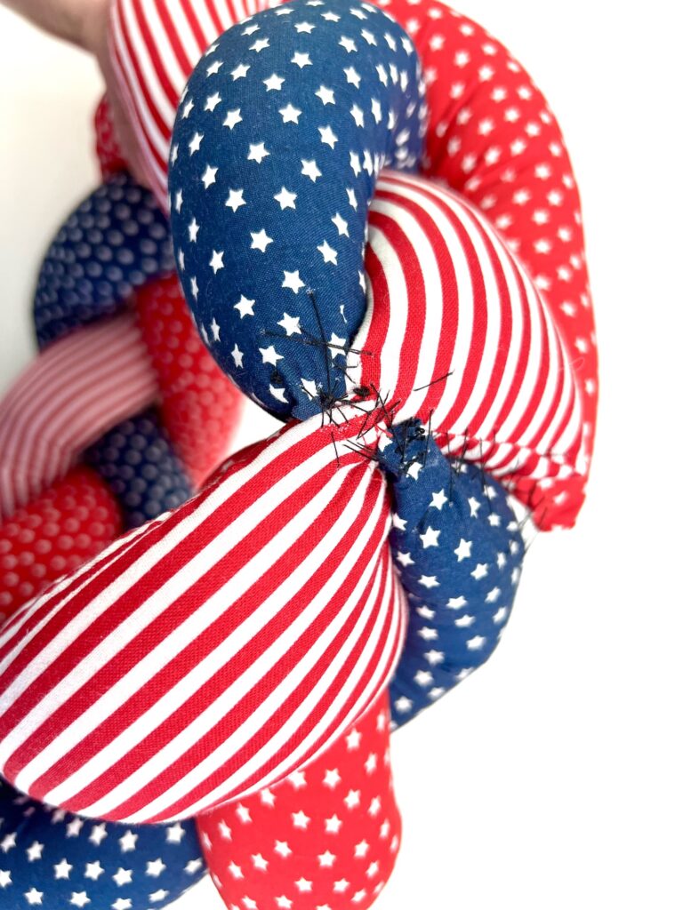A close-up of a braided fabric wreath with red, white, and blue striped and star-patterned sections, showing visible stitches at a seam.