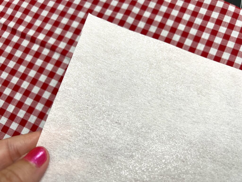 A hand with pink nail polish holds a white textured sheet over a red and white checkered fabric surface.